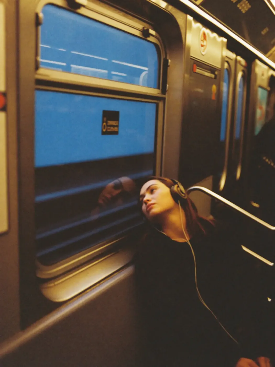 A woman with headphones leaning against a subway window